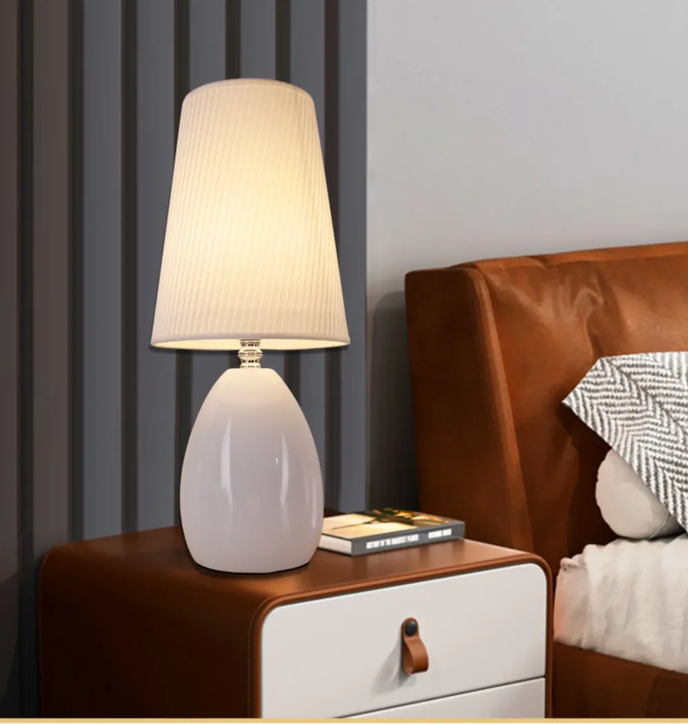 Đèn bàn chao vải trắng xếp ly, chân sứ trắng dáng trứng, đặt cạnh giường da nâu