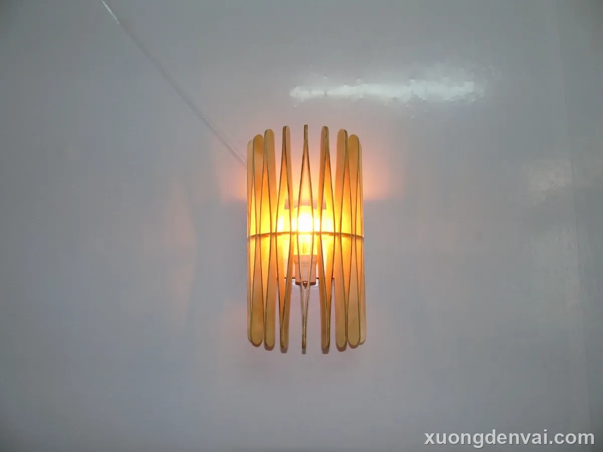 Đèn gỗ áp tường dáng phễu loe, nan gỗ cong xòe rộng dần từ điểm gắn tường xuống đáy, ánh sáng vàng ấm bên trong