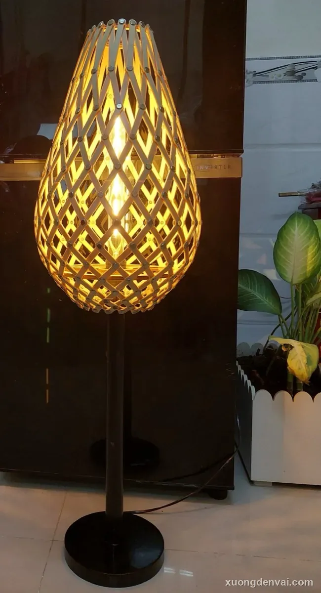 Đèn bàn nan gỗ đan chéo tạo lưới mắt cáo, thân đèn dáng giọt nước, đặt trên chân trụ đứng cạnh chậu cây và tủ gỗ tối màu