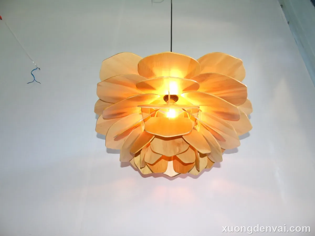 Đèn thả trần gỗ dáng hoa nở với nhiều lớp cánh gỗ xòe rộng, ánh sáng vàng ấm rực từ tâm đèn, nền tường trắng