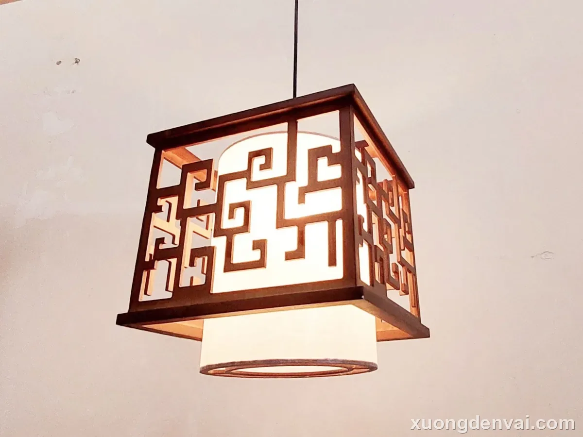 Đèn thả trần gỗ dạng khối vuông, khung gỗ tối màu chạm hoa văn hồi văn kiểu Á Đông, phía dưới có chao vải kem viền tròn