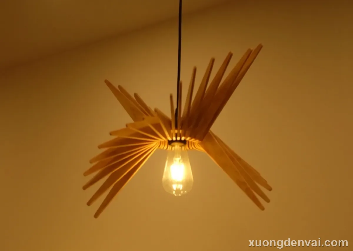 Đèn gỗ thả trần với các thanh nan gỗ mảnh tỏa ra bốn phía như đóa hoa nở, bóng đèn dây tóc sáng vàng treo giữa tâm