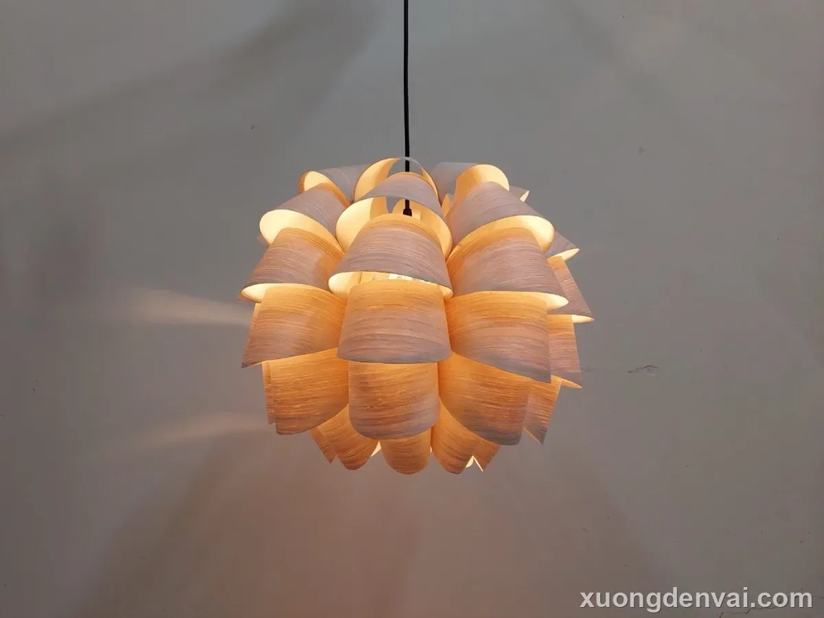 Đèn thả trần gỗ dáng quả thông với các vảy gỗ hình lục giác xếp lớp đều, ánh sáng vàng ấm lan tỏa qua từng khe hở, nền tối