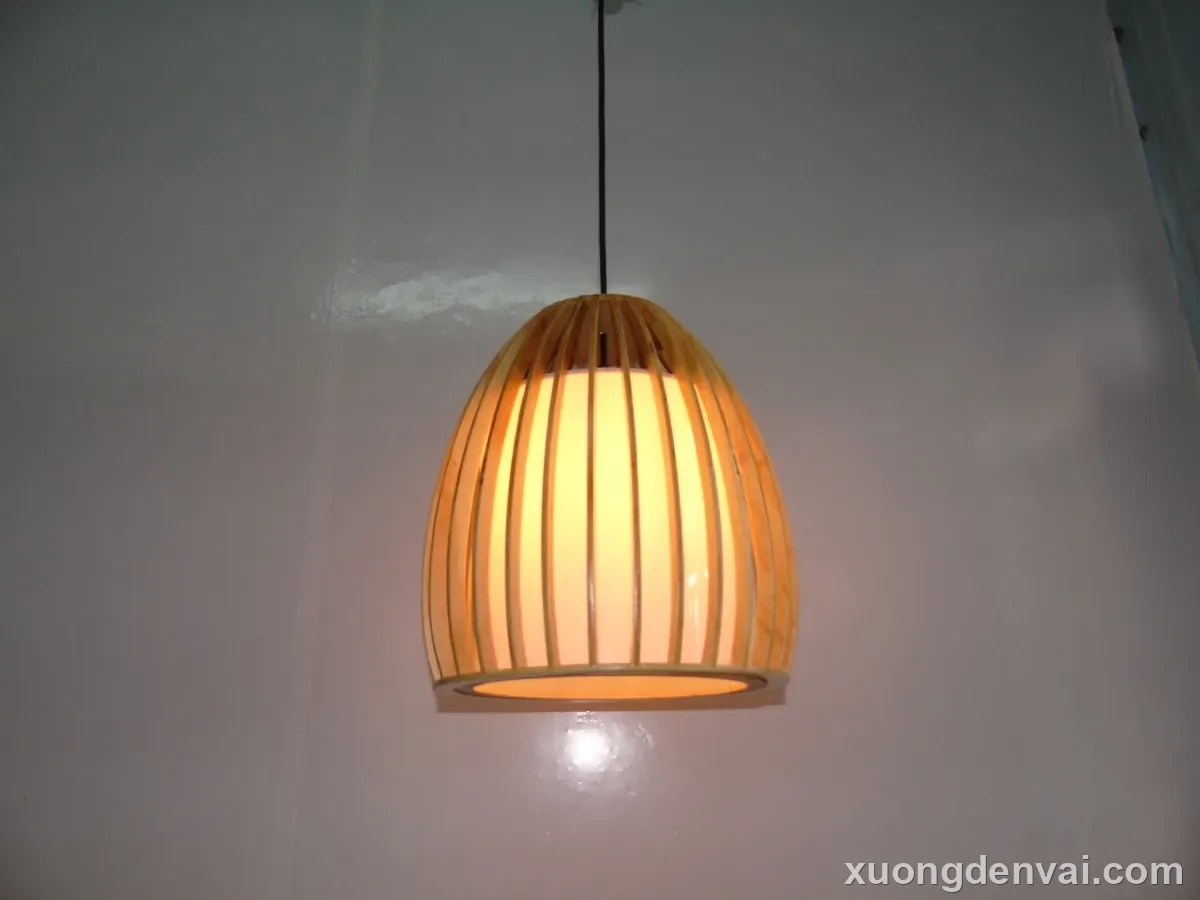 Đèn gỗ thả trần dáng vòm hình quả trứng đứng, các nan gỗ chạy dọc đều từ đỉnh xuống đáy, ánh sáng vàng ấm hắt ra ngoài