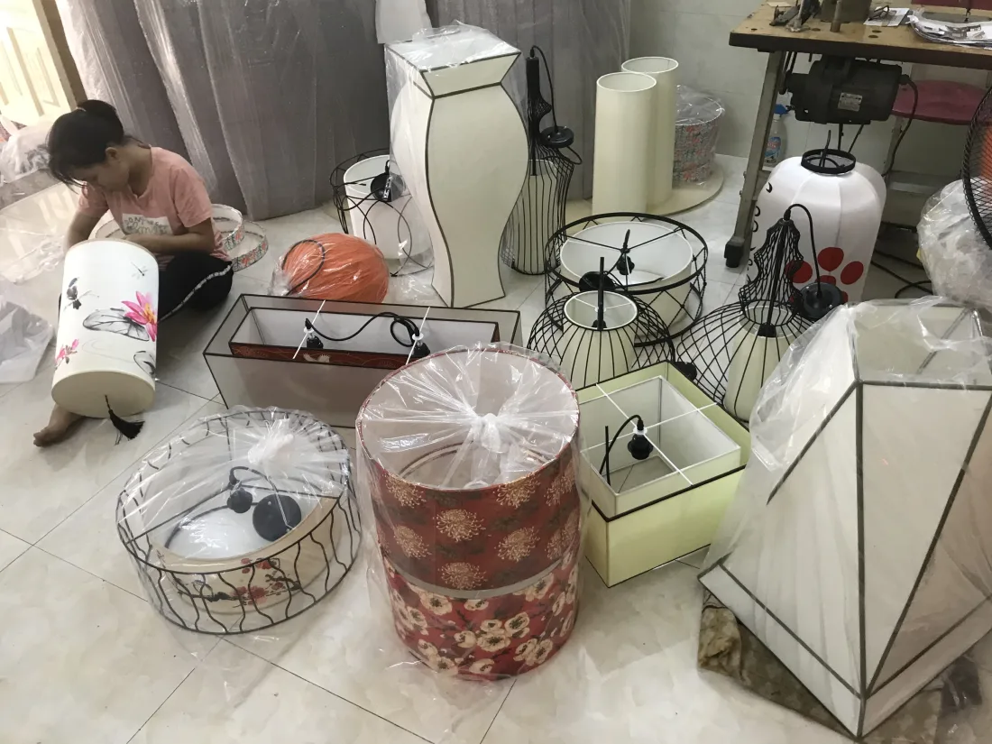 Góc xưởng gia công đèn vải: thợ đang hoàn thiện nhiều chao đèn vải đa dạng dáng — trống, vuông, lồng khung sắt, vẽ hoa sen — một số đã bọc nilon chờ giao